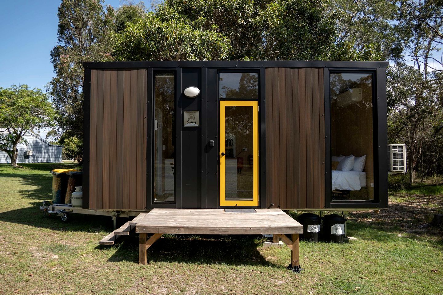 Eine kleine, moderne Hütte mit einer Holterrasse, einer Tür mit gelbem Rahmen und einem sichtbaren Bett darin.
