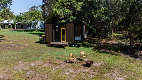 A small cabin with a wooden deck, a bench, and two chairs in a grassy area with trees.