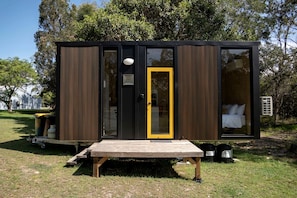 Uma cabana pequena e moderna, com deck de madeira, uma porta única com moldura amarela e uma cama visível no interior.