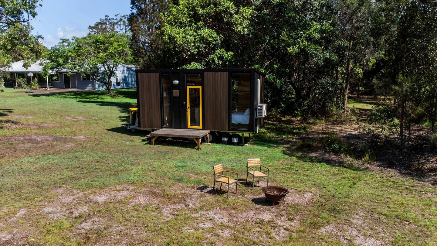 A small cabin with a wooden deck, a bench, and two chairs in a grassy area with trees.