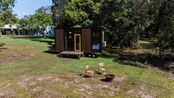 A small cabin with a wooden deck, a bench, and two chairs in a grassy area with trees.