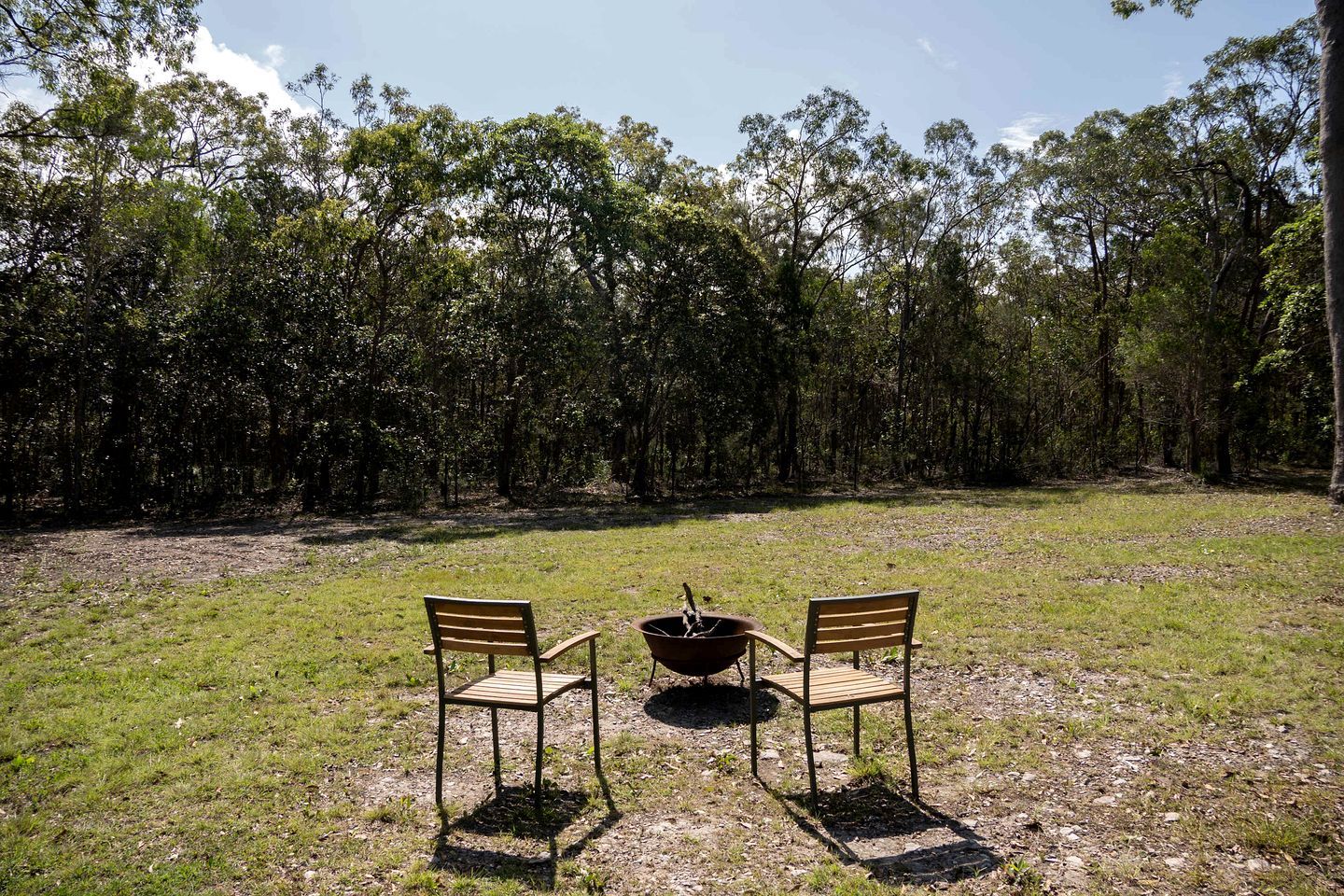 Un espace extérieur aménagé avec deux chaises en bois et une table en métal, entouré d’un champ herbeux et d’arbres denses.