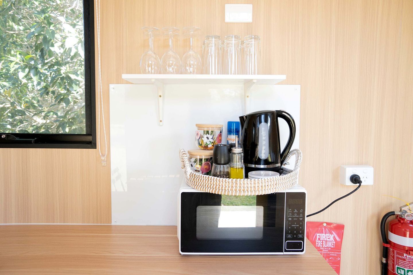 A kitchen with a microwave, a coffee maker, and a shelf with glasses and a fire extinguisher.