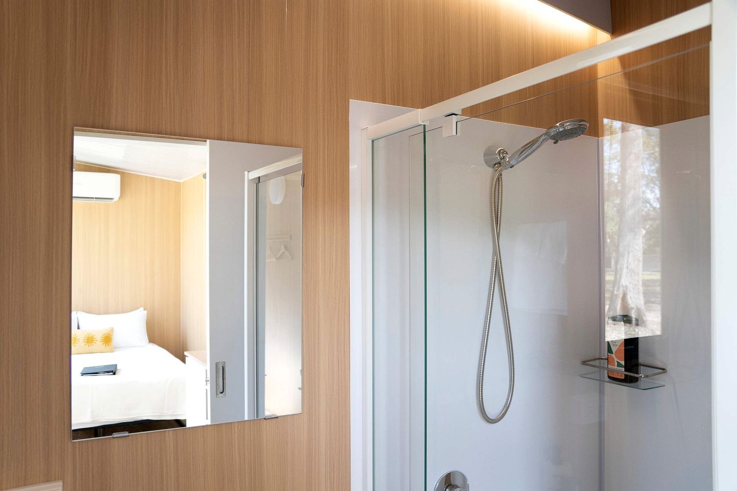 Un baño moderno con cabina de ducha de vidrio, un perchero visible, y una cama con cabecero blanco en la habitación contigua.
