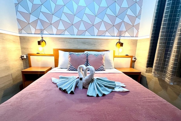 Habitación de hotel con cama, dos lámparas de noche, techo con estampado geométrico y una escultura de toalla con forma de cisne.