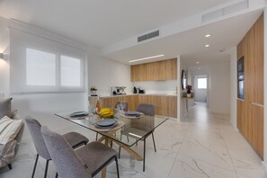 Eine moderne Küche mit Holzschränken, eine Essecke mit einem Glastisch und ein Wohnbereich mit einer Couch und Sesseln.