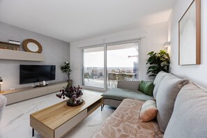 Ein modernes Wohnzimmer mit Flachbildfernseher, Couchtisch, Sofa und großem Fenster mit Blick auf Gebäude.