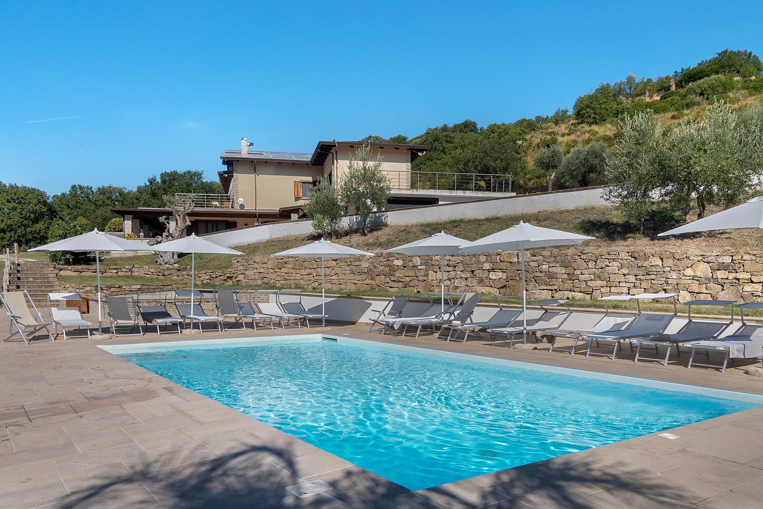 A swimming pool with lounge chairs and umbrellas, surrounded by a stone wall and a hillside with trees.