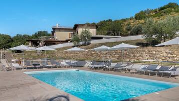 A swimming pool with lounge chairs and umbrellas, surrounded by a stone wall and a hillside with trees.