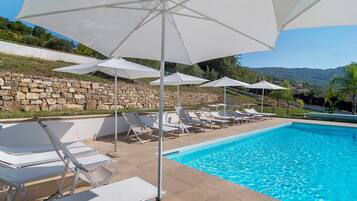 Una piscina con sillones blancos y sombrillas, rodeada de muros de piedra y follaje.