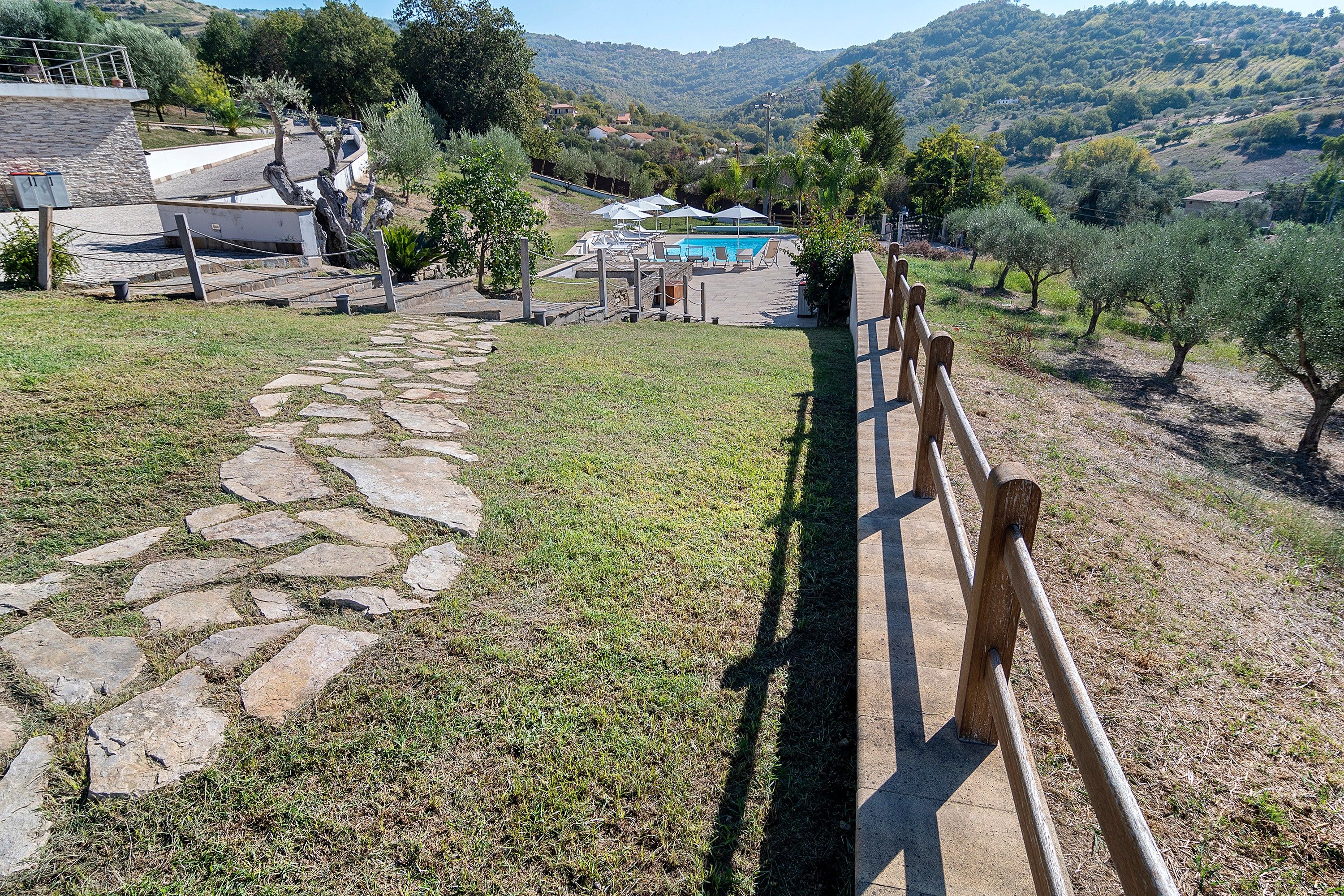 Ein Steinweg führt zu einem Poolbereich mit Holzzaun, umgeben von Grünflächen und Hügeln.