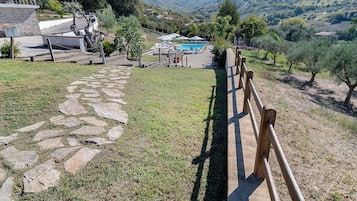 Ein Steinweg führt zu einem Poolbereich mit Holzzaun, umgeben von Grünflächen und Hügeln.