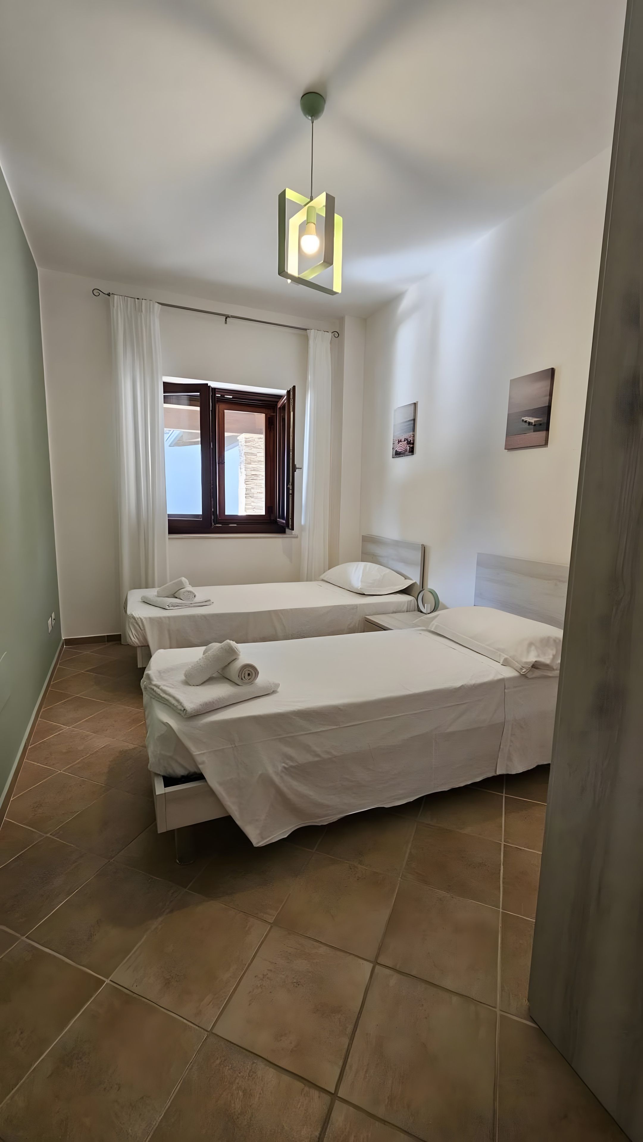 Une chambre d’hôtel avec deux lits, une fenêtre avec des rideaux, un luminaire au plafond et un tableau accroché au mur.