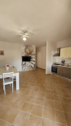 Een ruime woonkamer met een eettafel, stoelen, een plafondventilator, en een keuken met kastjes en een fornuis.