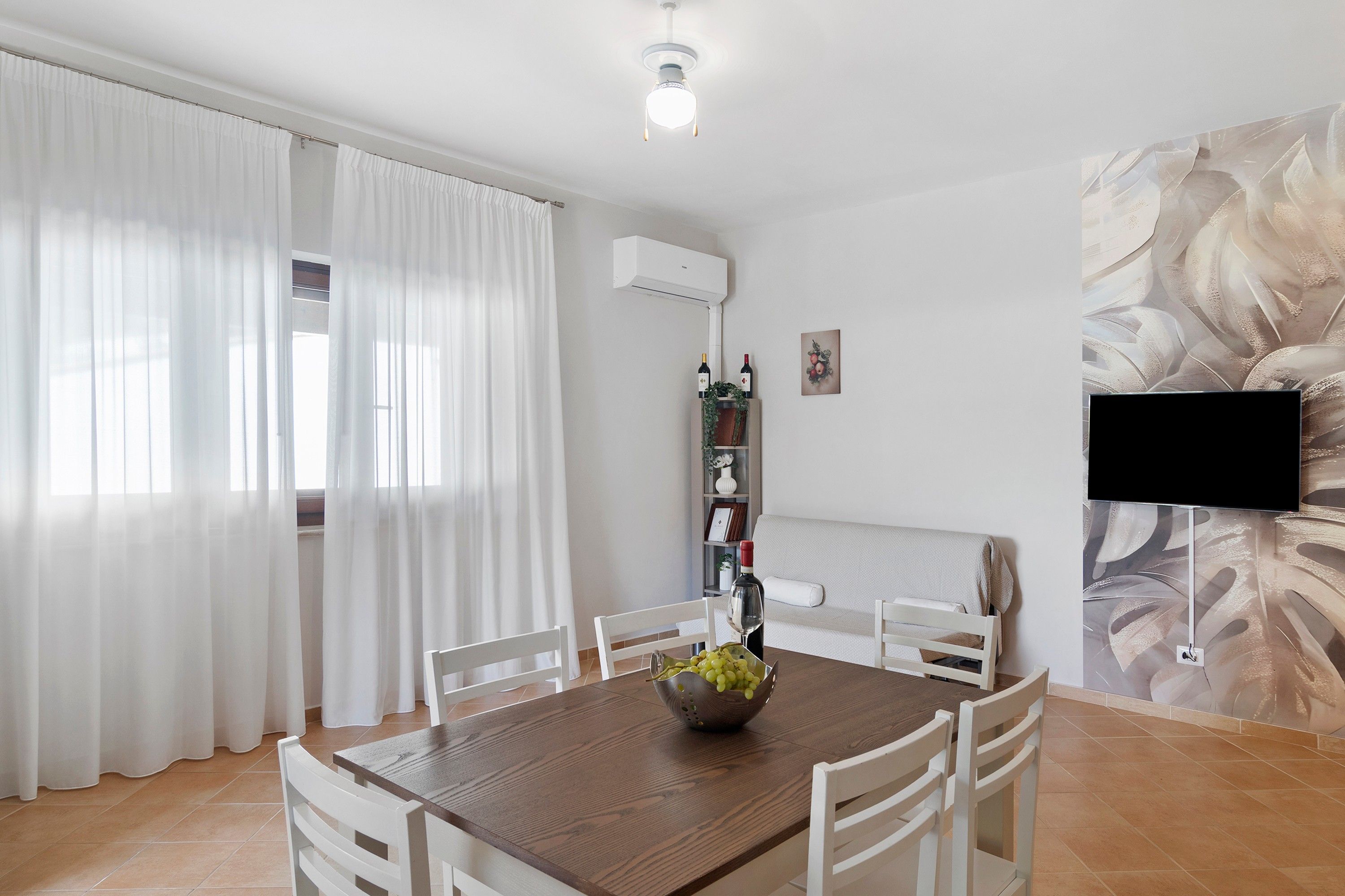 Un comedor con una mesa de madera, sillas blancas, un cuenco de uvas y una botella de vino.