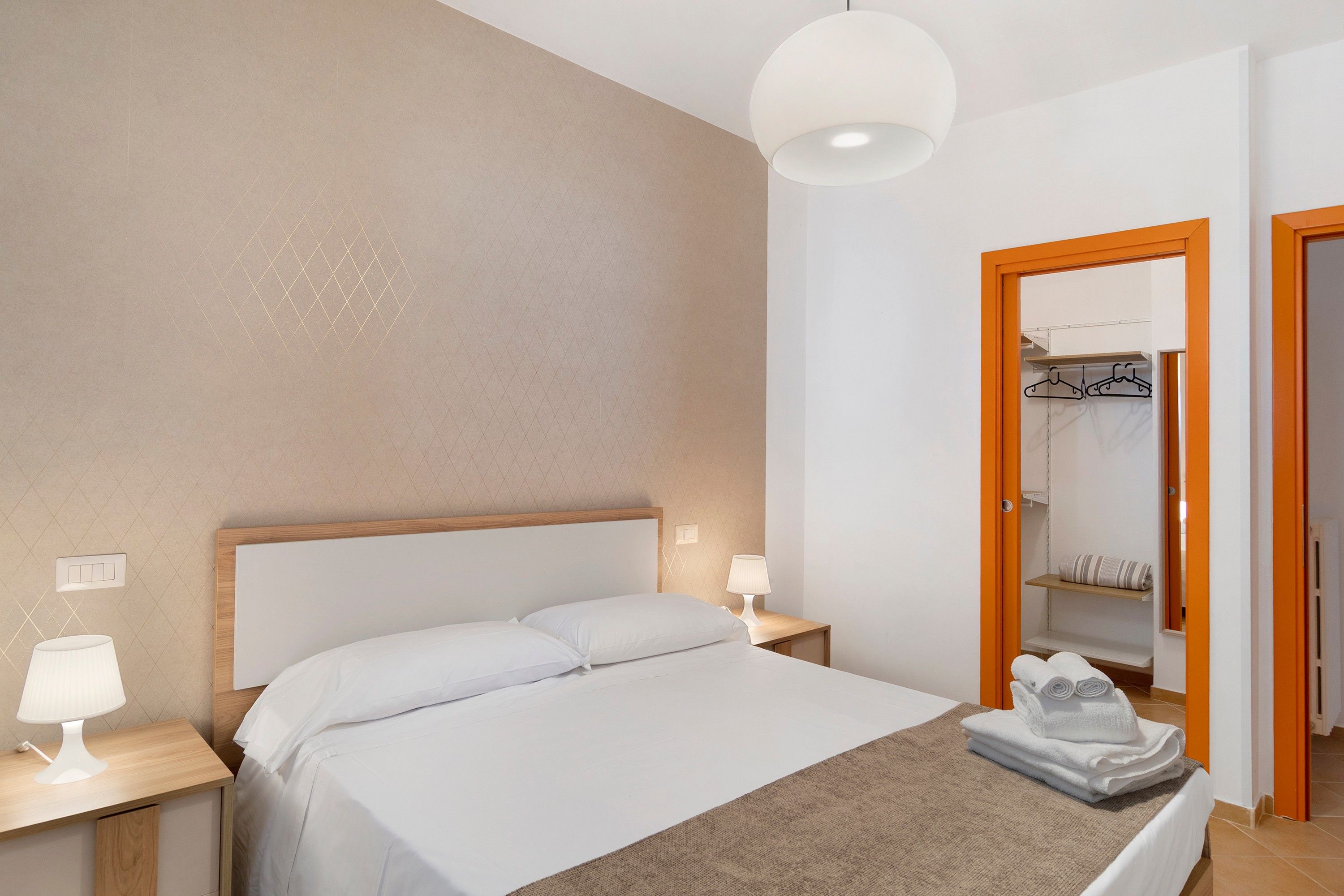 Hotellihuone, jossa on sänky, yöpöydällä olevat lamput, yöpöytä, pyyhe ja peilillä varustettu vaatekaappi.