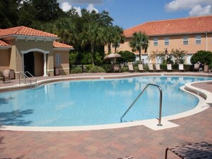 Une piscine avec une terrasse carrelée, des chaises longues et un bâtiment surmonté d’un toit recouvert de tuiles rouges.
