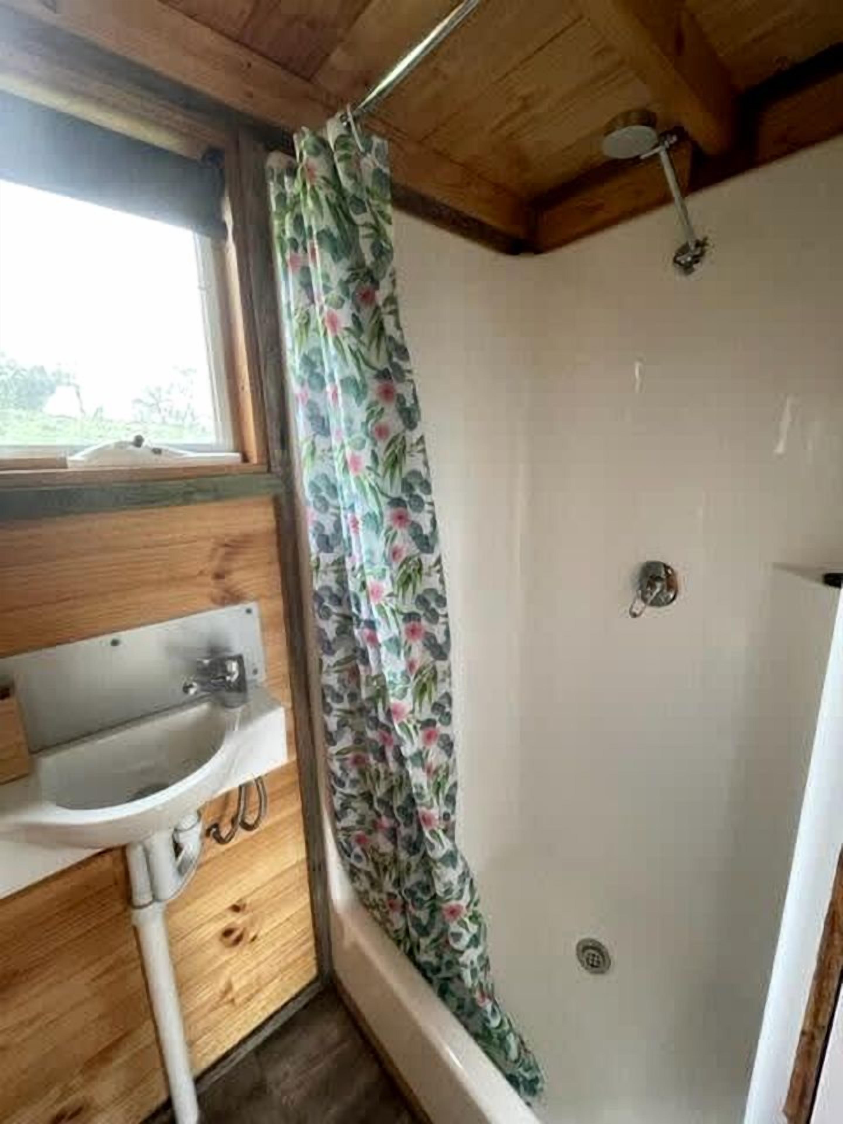 Une salle de bain avec un plafond en bois, un lavabo blanc, une douche avec un rideau à motifs floraux et une fenêtre.