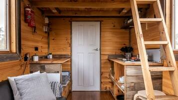 A compact cabin with a wooden interior, a white door, a small kitchenette, a black sofa with cushions, and a wooden ladder.