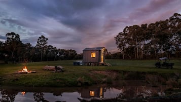 A small cabin with a porch, a fire pit, and a reflection in a body of water.