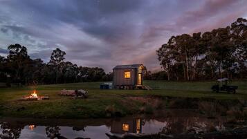 A small cabin with a porch, a fire pit, and a reflection in a body of water.