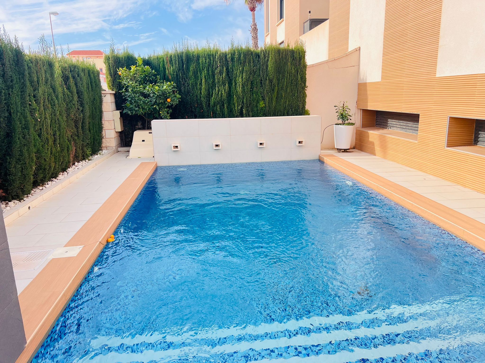 A rectangular swimming pool with a tiled deck, surrounded by a low hedge and a building with a beige facade.