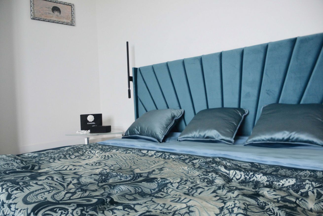 Une chambre à coucher avec un lit, des oreillers, une table de chevet sur laquelle se trouve un sac Chanel, et un tableau accroché au mur.