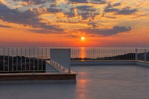 En takterrass med ett metallräcke, en trappa och utsikt över solnedgången över en vattenyta.