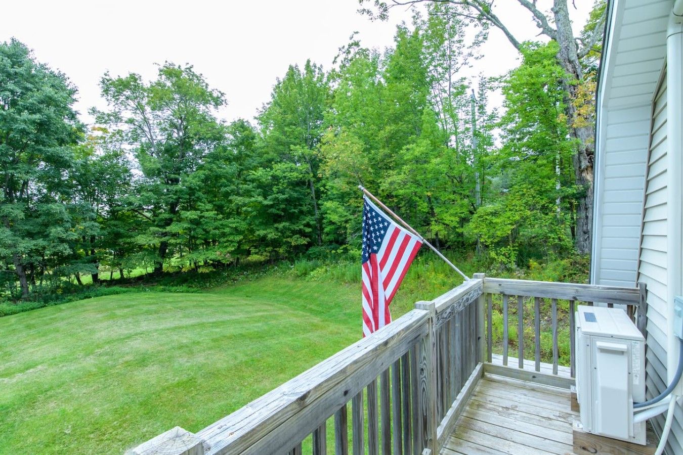 A wooden deck with a flag, surrounded by a well-maintained lawn and dense trees.