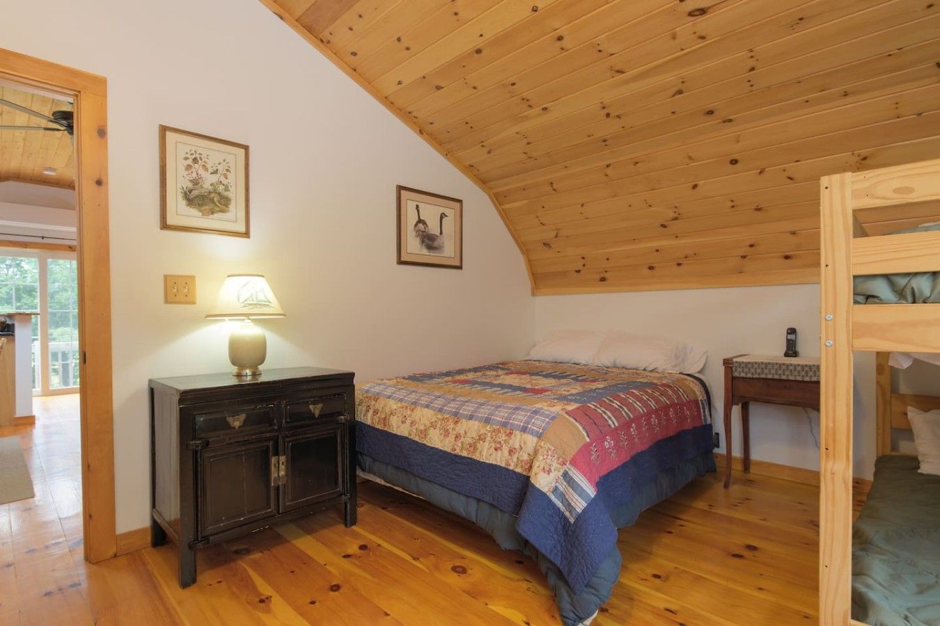 Une chambre avec un plafond en bois, un lit recouvert d’une couvre-lit à motifs, une table de chevet avec une lampe et un tableau encadré au mur.