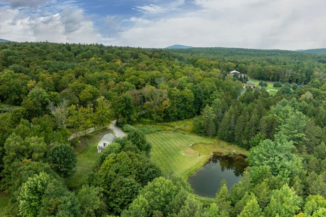 A rural landscape with a house surrounded by dense forest, a pond, and a golf course in the distance.