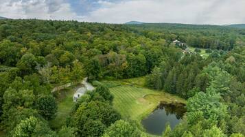 A rural landscape with a house surrounded by dense forest, a pond, and a golf course in the distance.
