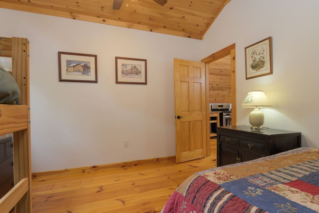 A bedroom with wooden flooring, a bed with a patterned blanket, a dresser, a nightstand with a lamp, and two framed pictures on the wall.