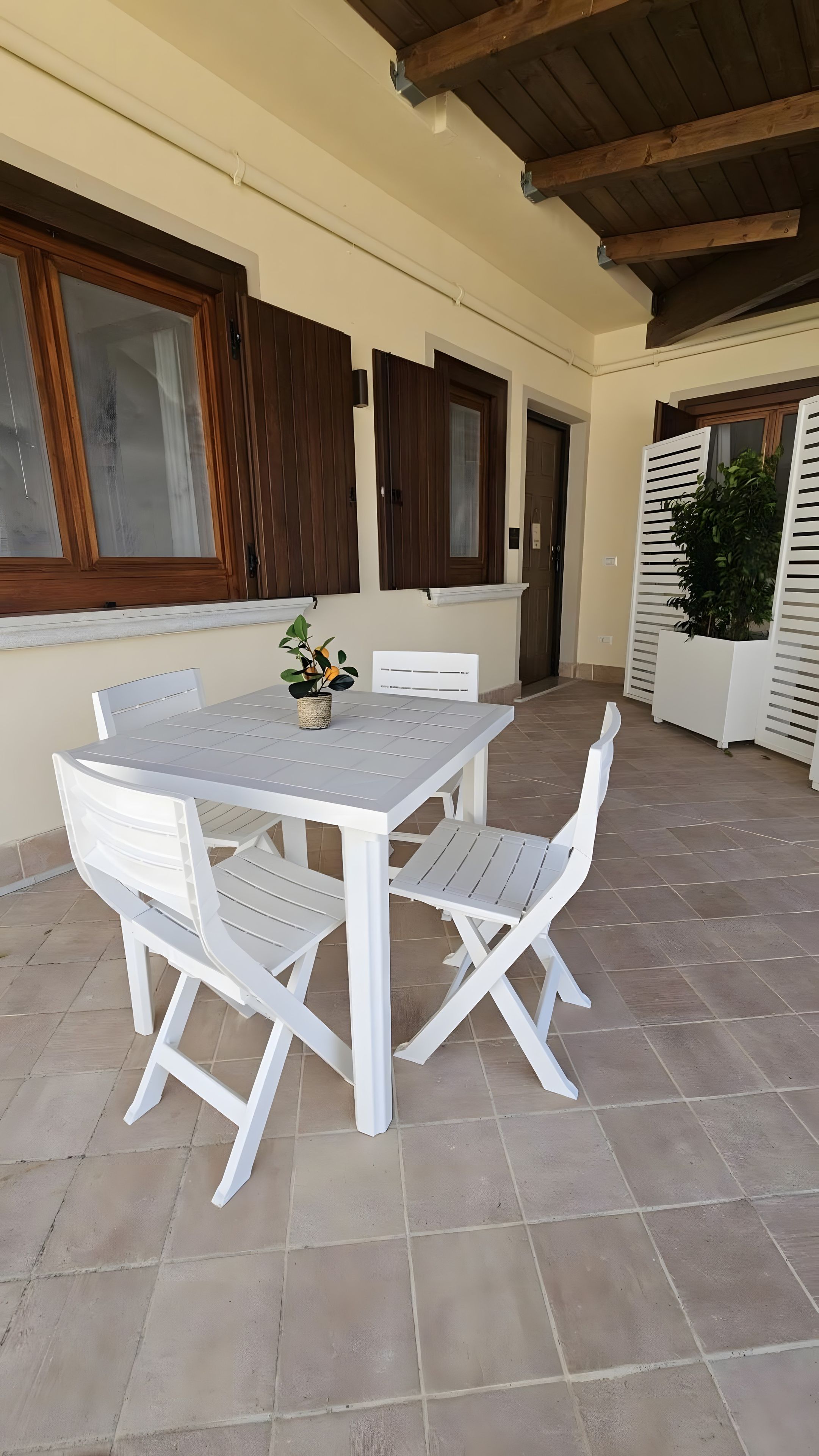Un patio al aire libre con una mesa y sillas blancas, una planta en maceta y un suelo de baldosas.