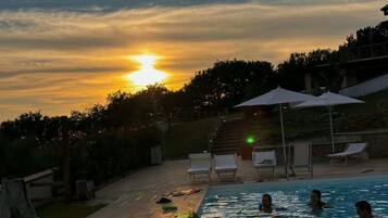 A swimming pool with people in the water, surrounded by lounge chairs and umbrellas, with a sunset in the background.