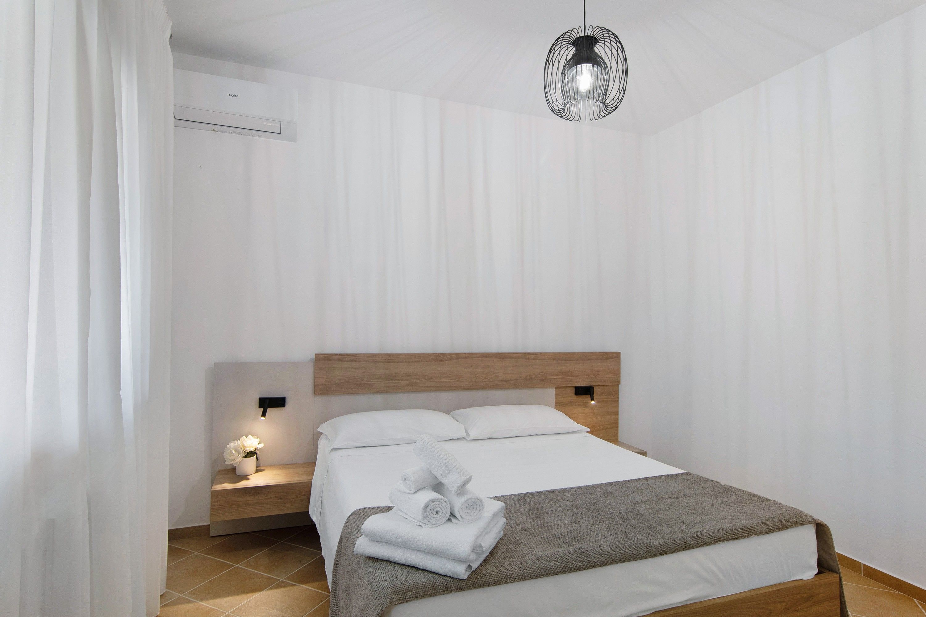 Et moderne hotelværelse med sengegavl i træ, en pænt redt seng med hvide sengetøj, et lille natbord med lampe og en hængende lampe.