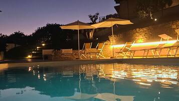 Ein Schwimmbecken mit Liegestühlen und Sonnenschirmen, umgeben von Steinmauern und Bäumen.