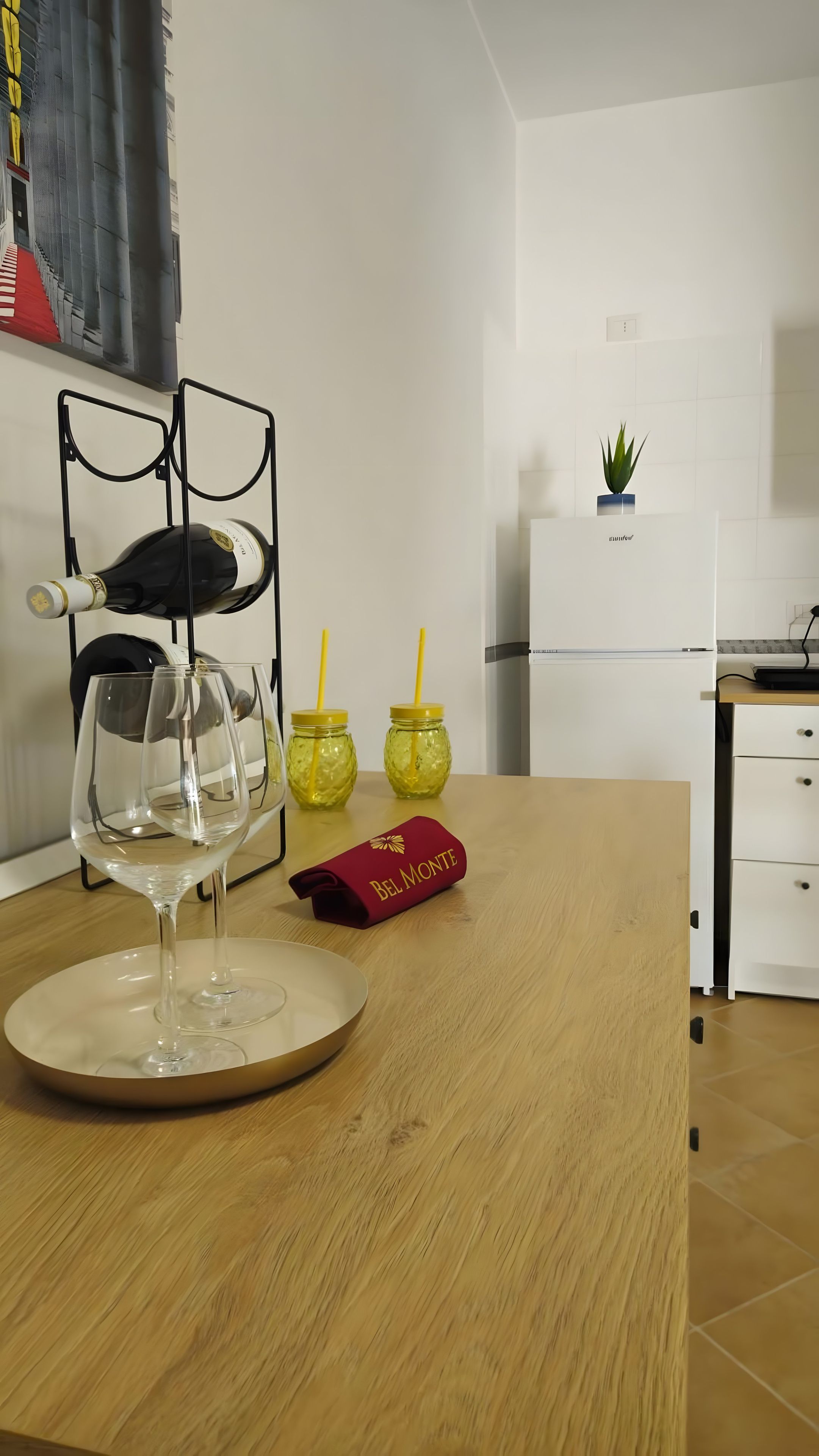 Une cuisine avec une table en bois, un verre à vin, deux bocaux en verre avec des couvercles jaunes et une boîte rouge portant l’inscription « BEL MONTE ».