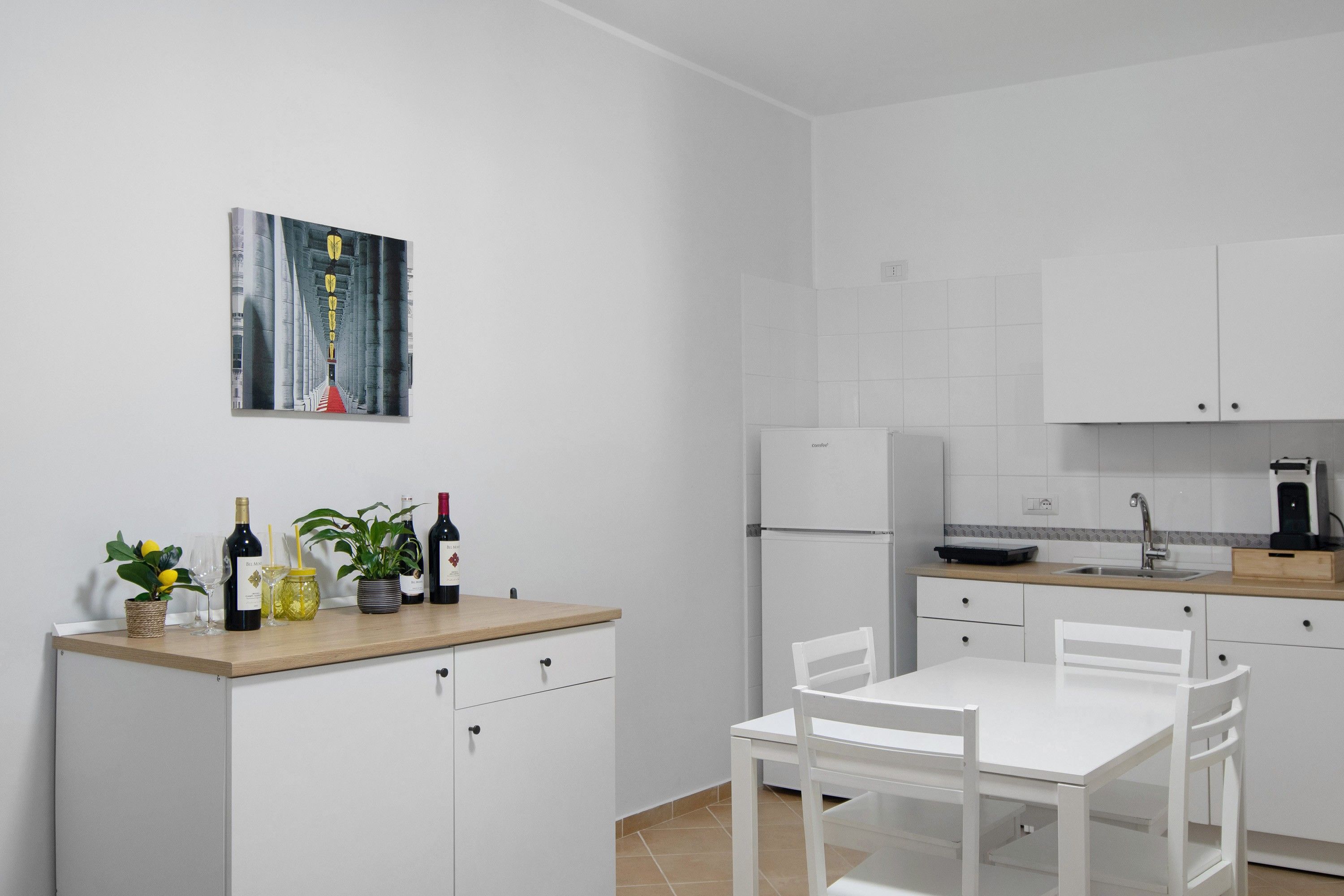 Una cocina moderna con armarios blancos, una mesa de comedor con sillas, una cafetera y una botella de vino sobre la encimera.