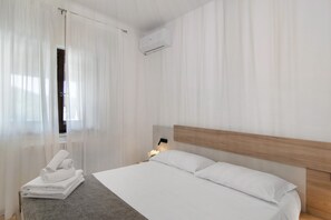 A hotel room with a bed, a nightstand with a lamp, a window with sheer curtains, and a wall-mounted air conditioner. - Apartment 'La Radice' with Mountain View, Wi-Fi and Air Conditioning (Agropoli)