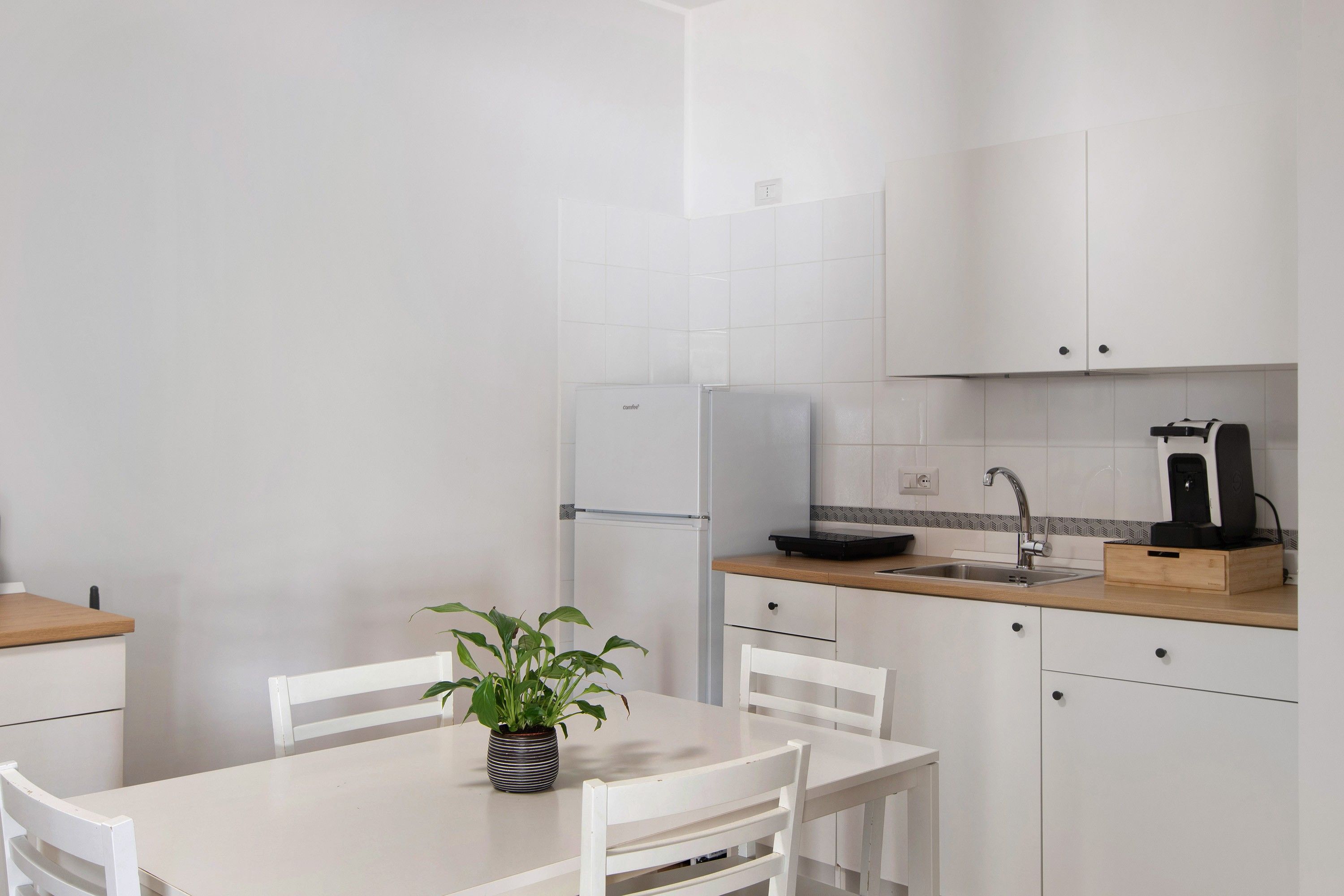 Una cocina moderna con muebles blancos, un refrigerador integrado, un lavamanos y una cafetera. Hay una mesa de comedor con sillas y una planta en maceta encima.