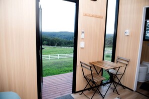 Een kamer met een houten tafel en stoelen, uitzicht op een groen veld met een witte omheining, en een houten deur.
