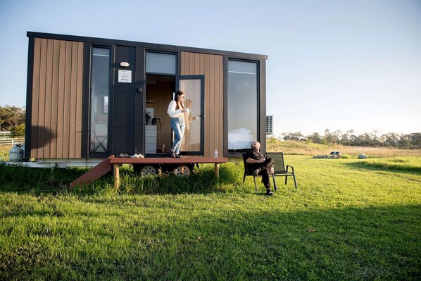 A modern, single-story building with a large sliding door, a small porch, and a grassy field in the background.