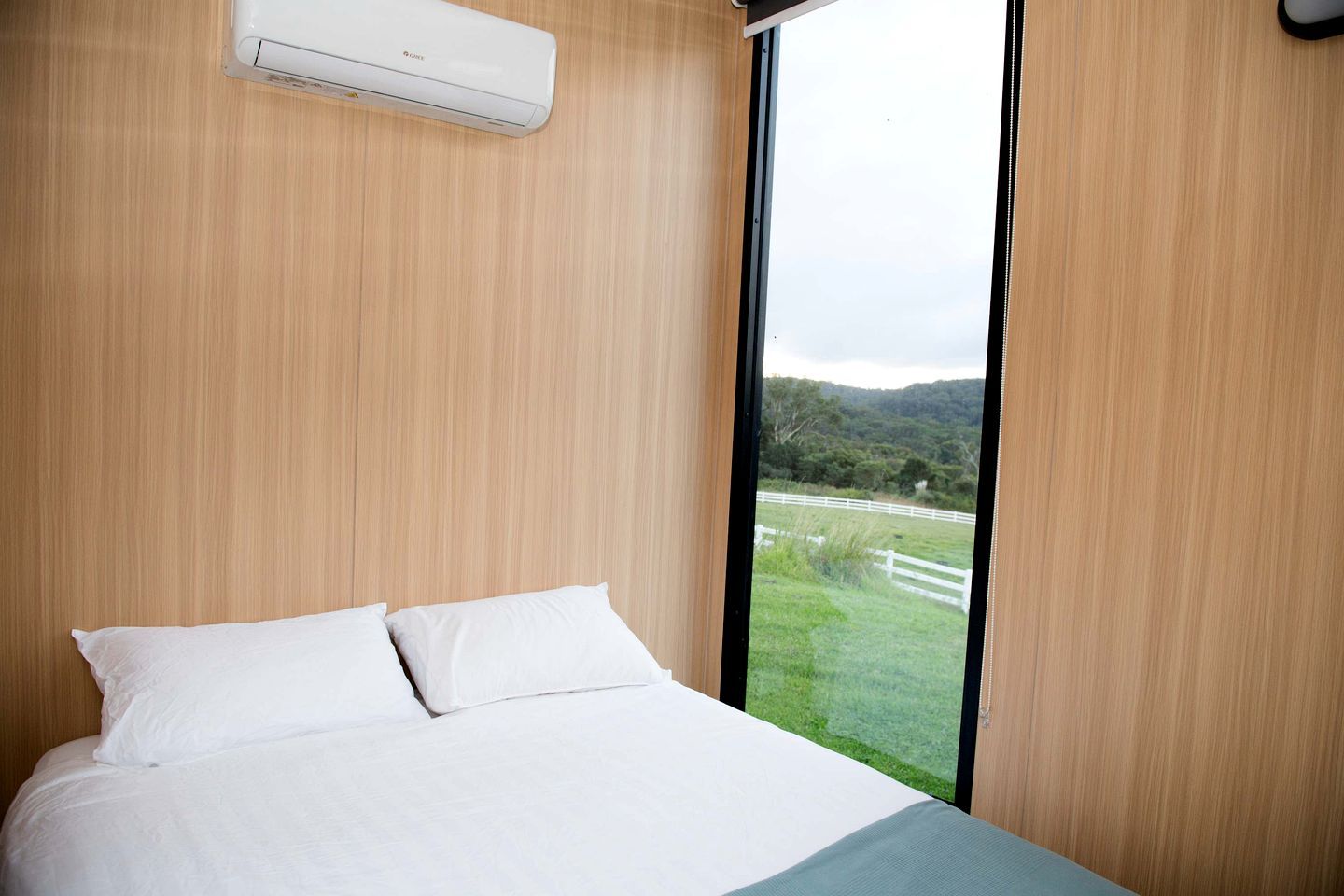Een slaapkamer met een bed, witte beddengoed, een raam met uitzicht op een groen landschap en een airconditioner aan de muur.