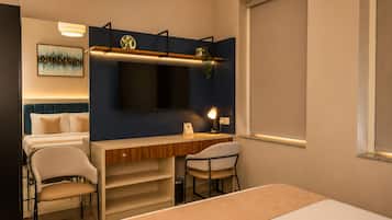 A hotel room with a bed, a desk, two chairs, a TV, and a painting on the wall.