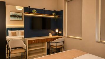 A hotel room with a bed, a desk, two chairs, a TV, and a painting on the wall.