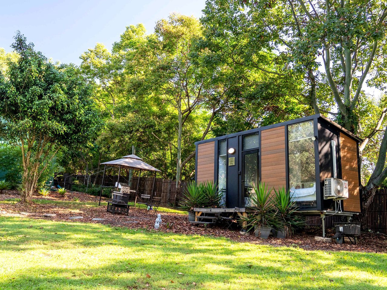 A modern, compact outdoor structure with a door and windows, surrounded by lush greenery and a patio area with a table and chairs.