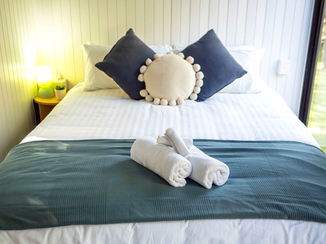 A neatly made bed with pillows and rolled towels, a bedside lamp, and a window with daylight.