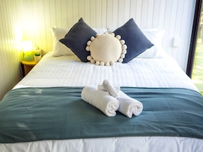 A neatly made bed with pillows and rolled towels, a bedside lamp, and a window with daylight.