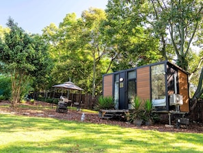 A modern, compact outdoor structure with a door and windows, surrounded by lush greenery and a patio area with a table and chairs.